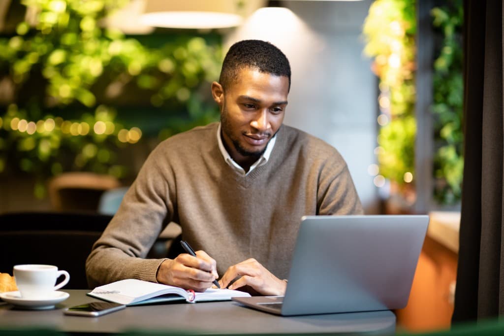 A professional Black man working on a laptop and writing in a notebook in a modern, sunlit cafe with greenery in the background