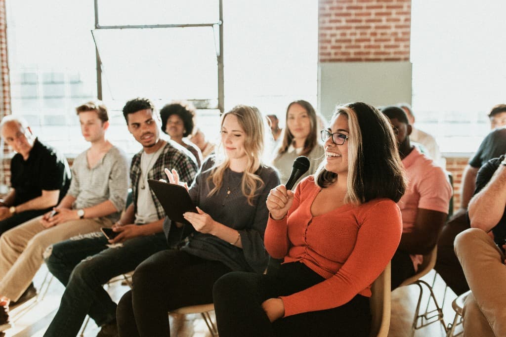 A diverse group of people participating in a workshop, with a woman in an orange top smiling and speaking into a microphone