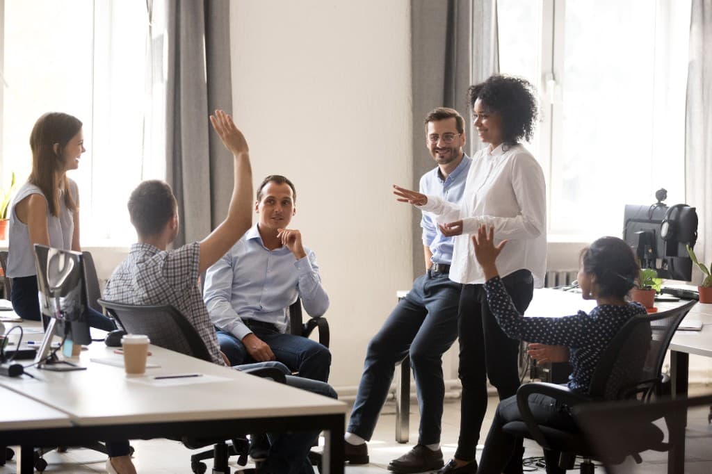 A diverse team in a bright modern office during a collaborative discussion, with team members raising hands as a woman stands and speaks to the group