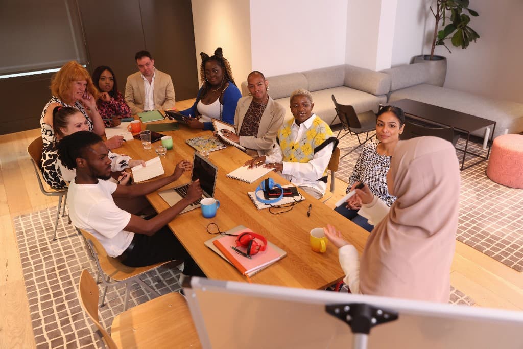 A diverse team in a modern office workshop, with a woman in a hijab standing and facilitating discussion around a conference table