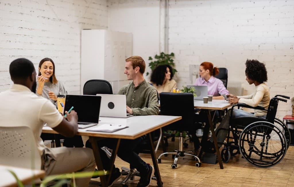 Colleagues collaborating in a bright open-plan office, including team members at shared tables with laptops in an inclusive workspace