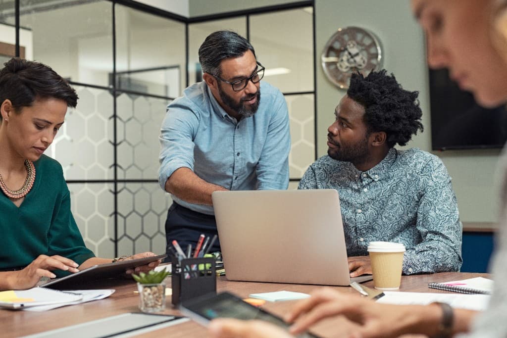 Colleagues collaborating around a conference table in a modern office, with one person standing to review a laptop while others participate with a tablet and notes