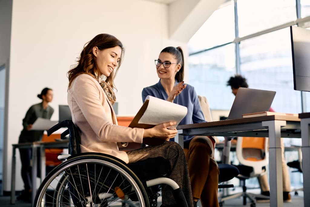 Two professional women collaborating in a bright, modern office. One woman sits in a wheelchair holding a clipboard, while her colleague at a desk gestures toward the document. They are both smiling in an inclusive, well-lit workspace