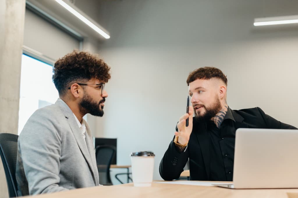 Two professional men in a modern office having a focused business discussion at a desk with a laptop and coffee